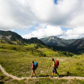 Two hikers walk on a winding trail through a vast, green mountain landscape under a partly cloudy sky.