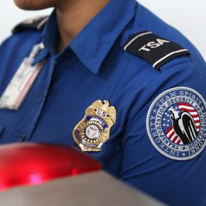 Security officer stands in blue TSA uniform, displaying badges on shoulder and chest, next to a glowing red-screened device.
