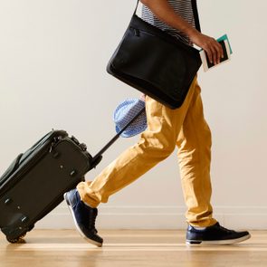 Man walks pulling suitcase, carrying shoulder bag and holding a book and passport, wearing yellow pants and blue sneakers in a minimal indoor setting.