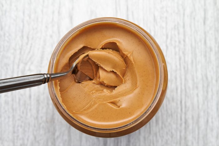A spoon scoops smooth peanut butter from a jar on a light wooden surface.