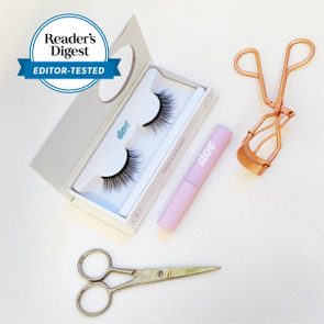 False eyelashes rest inside a box; nearby are scissors, a pink tube labeled 