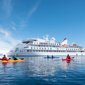 Kayakers paddle on calm water near a large white cruise ship in an icy, mountainous region under a clear blue sky.