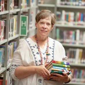 Person holds stack of books in a library aisle, surrounded by shelves filled with colorful books and green lettered signs.