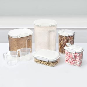 Plastic storage containers hold different foods, including sugar, nuts, and marshmallows, arranged on a white surface. Measuring cups are placed nearby.