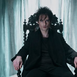 Person sitting on an ornate chair, looking intently forward, in a dimly lit room with sheer curtains.