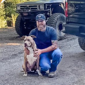Man crouches beside brown dog, surrounded by parked trucks and trees, in a wooded area.