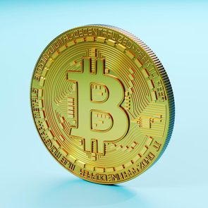 A gold Bitcoin coin stands upright against a light blue background, displaying a circuitry pattern and the Bitcoin symbol in the center.