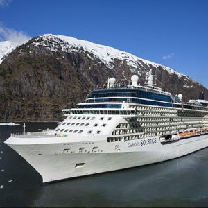 Cruise ship sails near snowy mountainous coast under clear blue skies, featuring multiple decks and lifeboats. 