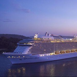 Cruise ship sails on calm water at dusk, illuminated by lights, against a backdrop of wooded hills and a twilight sky. Text reads 