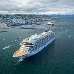 Cruise ship sails through a harbor surrounded by smaller boats, with a coastal city and snow-capped mountains in the background.