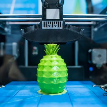 3D printer fabricating a green, geometric pineapple; set in a modern workshop with blue surfaces.