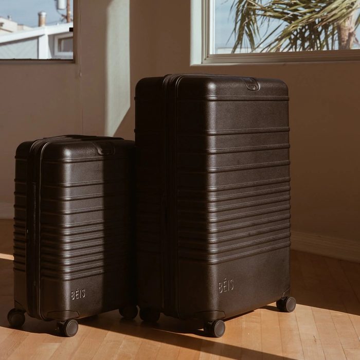 Two black suitcases stand on a wooden floor, sunlight streaming through a window with visible palm leaves outside.