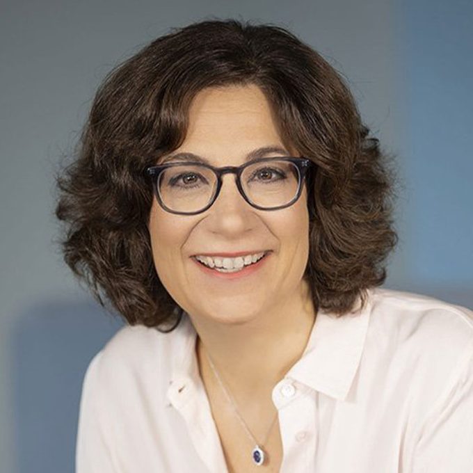 A person smiles warmly, wearing glasses and a white shirt, in front of a blurred blue background.