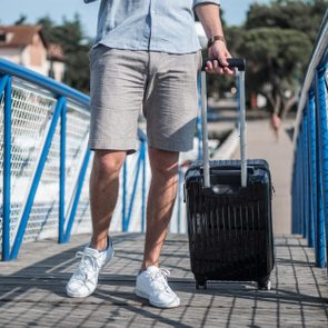 Person pulls a black suitcase across a blue-railed bridge on a sunny day, wearing casual summer clothes.