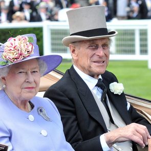 Two elegantly dressed individuals sit in a carriage at an outdoor event, surrounded by a lush green field and distant blurred spectators.