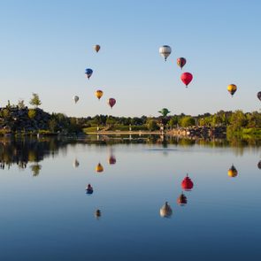 Hot air balloons float above a calm lake, reflecting in the water, surrounded by green trees and clear blue sky.