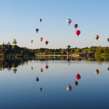 Hot air balloons float above a calm lake, reflecting in the water, surrounded by green trees and clear blue sky.