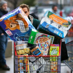 Shopping cart overflows with groceries, including paper towels and cereals, outside a store on a busy street. Shoppers and carts in the background.