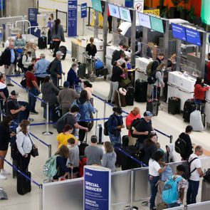 Travelers stand in line checking luggage at an airport counter, surrounded by signs and barriers in a busy terminal area.