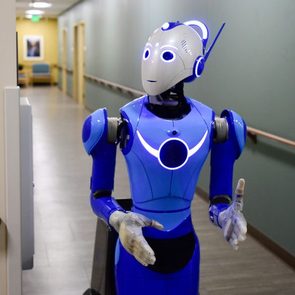A humanoid robot walks down a hospital hallway, surrounded by green walls and wooden railings, conveying a futuristic healthcare environment.