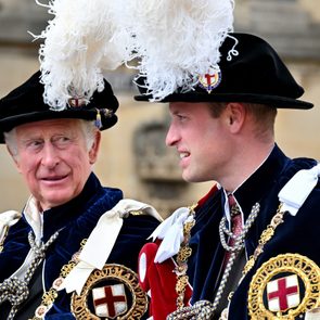 Two men in ornate blue velvet robes converse, wearing black hats adorned with white feathers, seated outdoors against a historic stone backdrop.