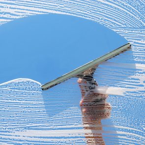 A squeegee wipes soap from a glass window against a clear blue sky, leaving streaks of foam and a clean surface behind.