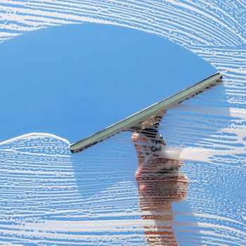A squeegee wipes soap from a glass window against a clear blue sky, leaving streaks of foam and a clean surface behind.