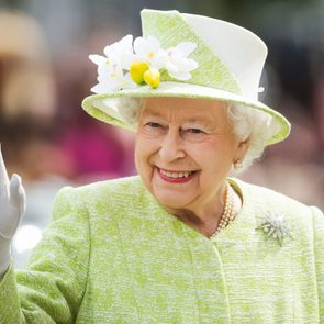 Woman waves, smiling warmly, wearing a bright green outfit with a floral hat. Blurred outdoor setting with people in the background.