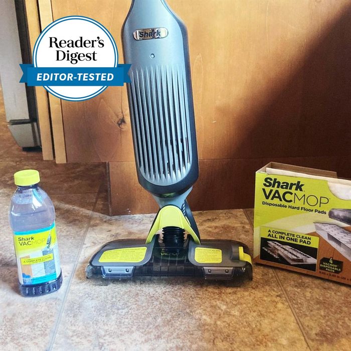 Shark VACMOP stands upright on a tiled floor, surrounded by cleaner bottle and disposable pad box, next to a wooden cabinet.