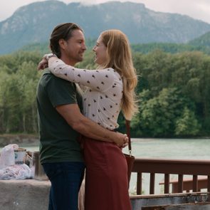 Couple hugging on a deck, surrounded by trees and mountains. Nearby, a table holds paint cans and cloths.