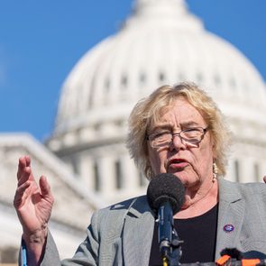 A person speaks passionately into a microphone outdoors, with a large white-domed building in the background under a clear blue sky.