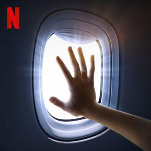 Hand reaching towards airplane window, illuminated by bright light; Netflix logo in the upper corner, set against a dark background.