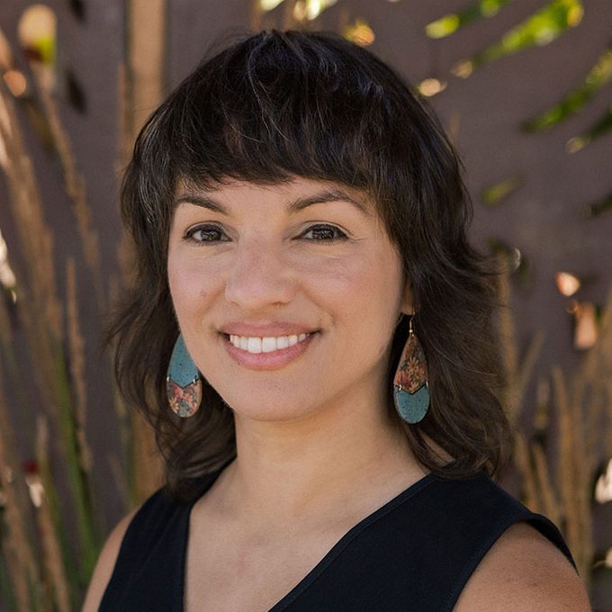 A woman smiles, wearing turquoise earrings with a nature-inspired pattern, against a blurred outdoor background with plants.