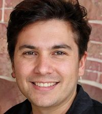 A person smiles while facing the camera, standing in front of a brick wall, wearing a black shirt, with dark hair.
