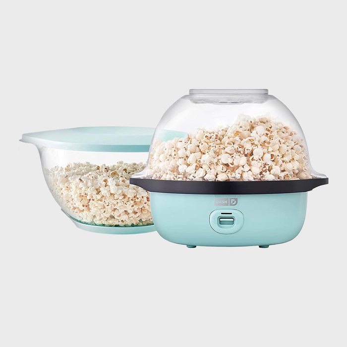 Popcorn maker pops corn, surrounded by a clear bowl filled with popcorn. The setting is neutral with a light grey background.
