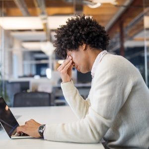 Person, sitting with hand on face in frustration, using laptop at a white desk in a modern office with glass walls and plants.