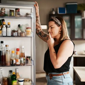A person examines refrigerator contents, hand on face, in a kitchen with dark cabinets, considering options among various bottles and jars.