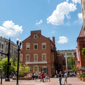 Brick building stands prominently; people walk and gather around, enjoying a sunny day in a plaza with streetlamps and shops. Sign reads 