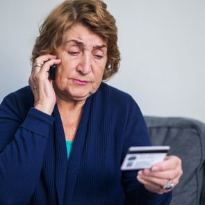 Woman holding phone and credit card, seated on a gray sofa, appears to be in a domestic setting.