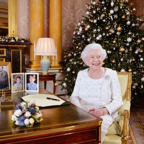 An elderly woman sits smiling at a decorated desk, a Christmas tree behind her in an elegant, warmly lit room.