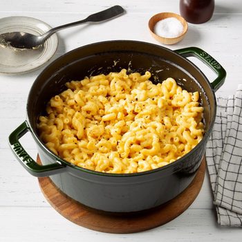 Macaroni sits in a pot on a wooden board, surrounded by salt bowl, spoon, and napkin on a white table.