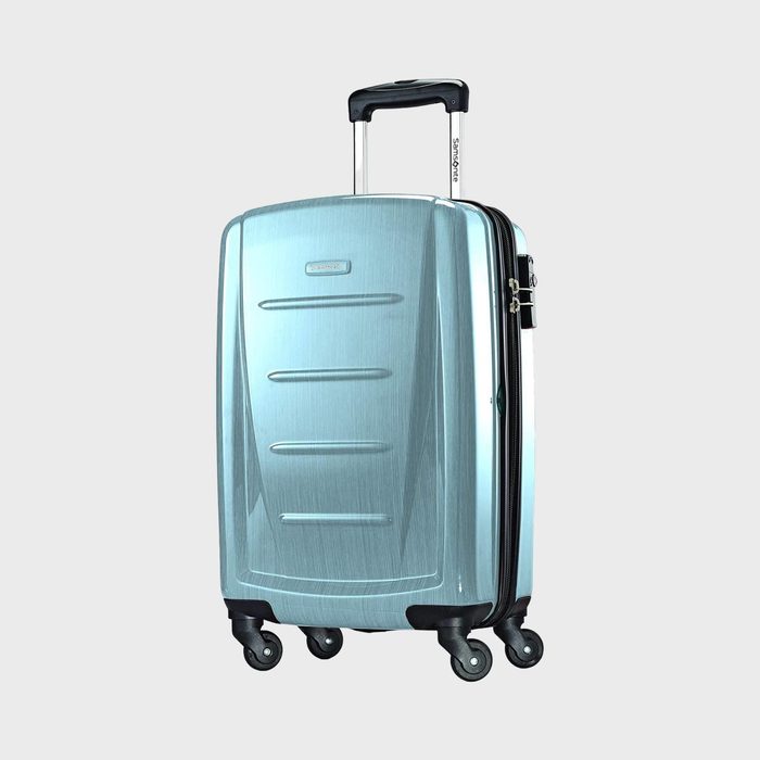 A metallic blue suitcase stands upright with an extended handle, featuring four double-spinner wheels, set against a plain grey background.