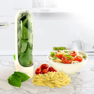 Container holds fresh spinach upright, next to uncooked pasta and a bowl of tomato pasta salad on a marble kitchen counter.