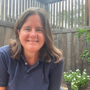 A person smiles, wearing a navy polo, seated outdoors with a wooden fence backdrop and potted white flowers nearby.