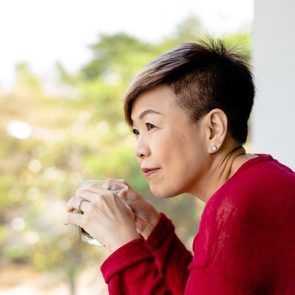A person holds a transparent cup, gazing thoughtfully, while wearing a red sweater in a bright, blurred outdoor setting.