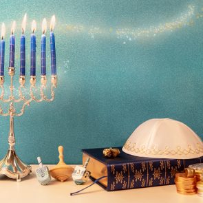 Menorah with lit blue candles stands; dreidels, coins, and kippah rest nearby on a book against a teal background with star accents.