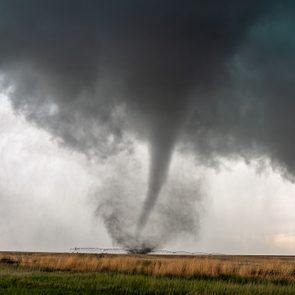 A tornado spirals violently, touching down on a flat, grassy landscape beneath dark, ominous clouds.