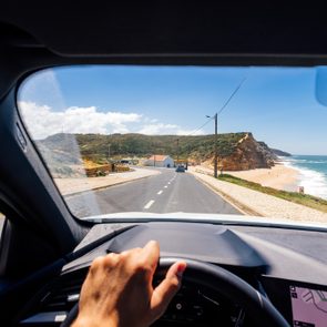 Hand grips steering wheel, driving along coastal road, blue sea and cliffs visible through windshield in sunny weather.