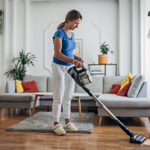 A person vacuums the living room floor; surrounded by a sofa, colorful cushions, and houseplants.