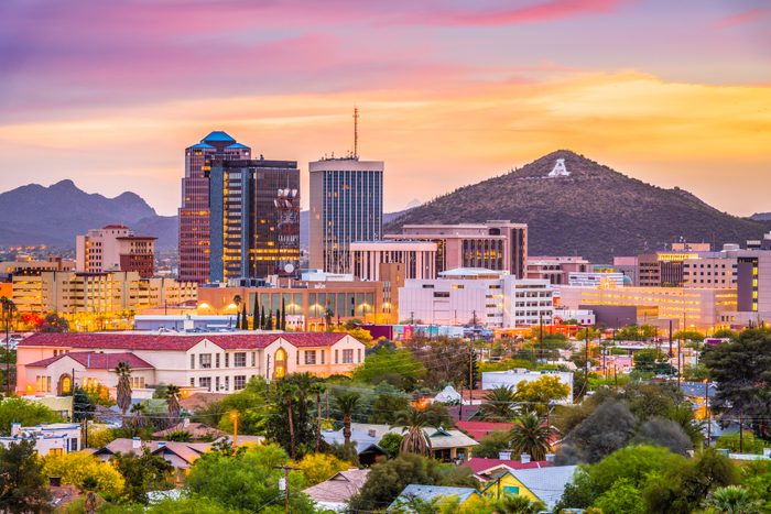 City skyline with tall buildings stands against a pink-orange sunset, surrounded by lush greenery and distant mountains in the background.